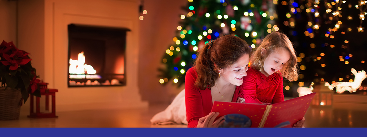 Mother and daughter read a book at fireplace on Christmas eve. Family with child celebrating Xmas. Decorated living room with tree, fire place and candles. Winter evening at home for parents and kids.